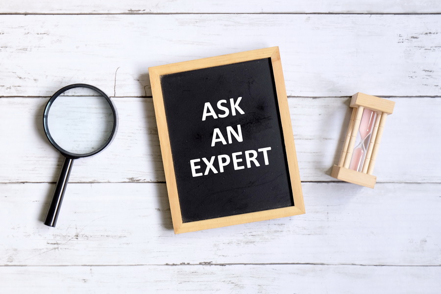 Top view of magnifying glass,hourglass and blackboard written with 'ASK AN EXPERT' on white wooden background.
