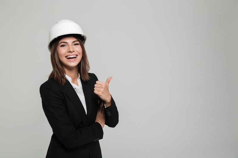Portrait of a cheerful pretty woman in hard hat and suit looking at camera and showing thumbs up gesture with copy space isolated over white background